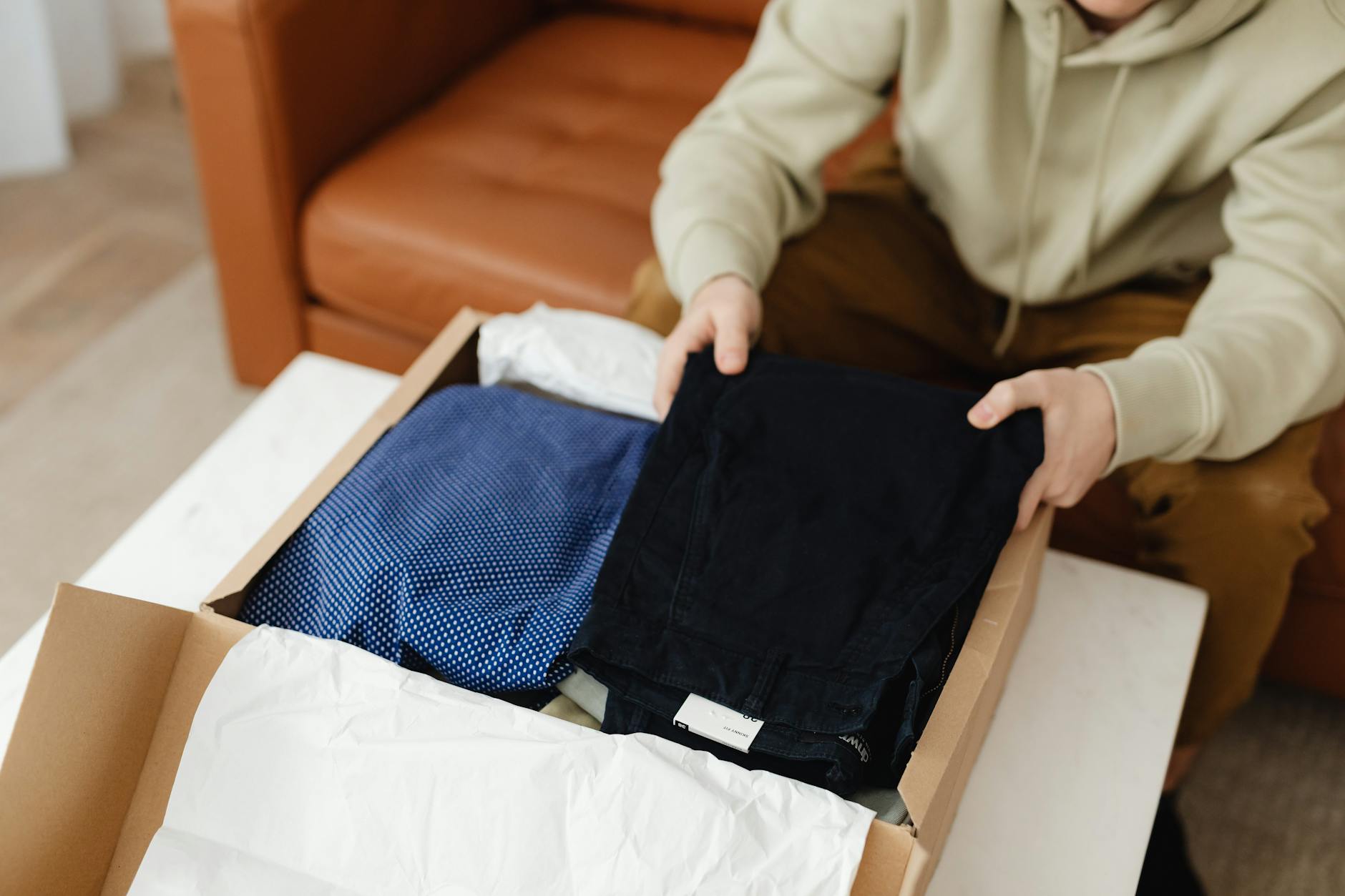 open clothing delivery box on a living room rug with neatly arranged outfits, a style card, and a person holding a phone reviewing items