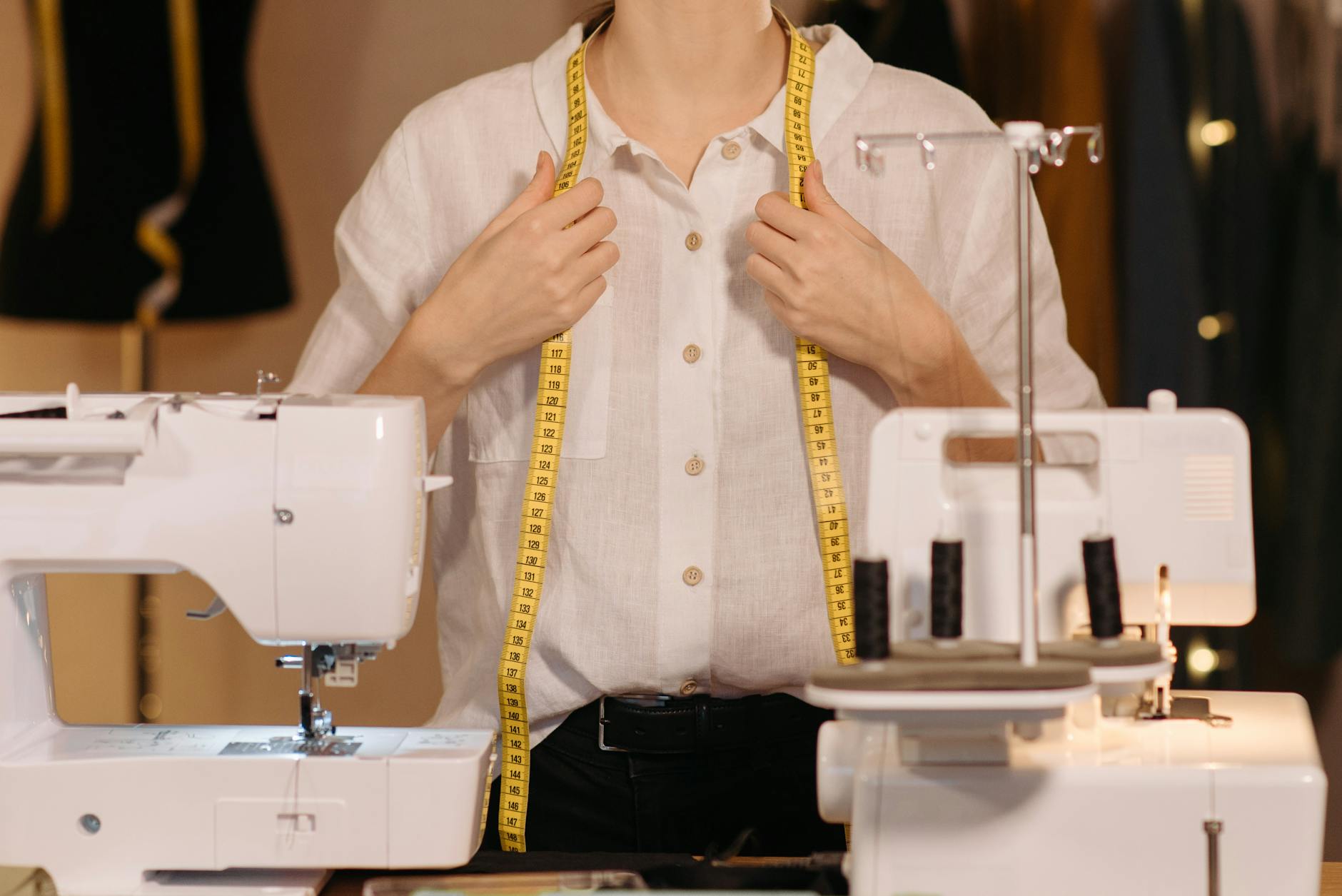shopper holding a smartphone showing a clothing app product page, with neatly folded shirts and a measuring tape on a bright bedroom dresser